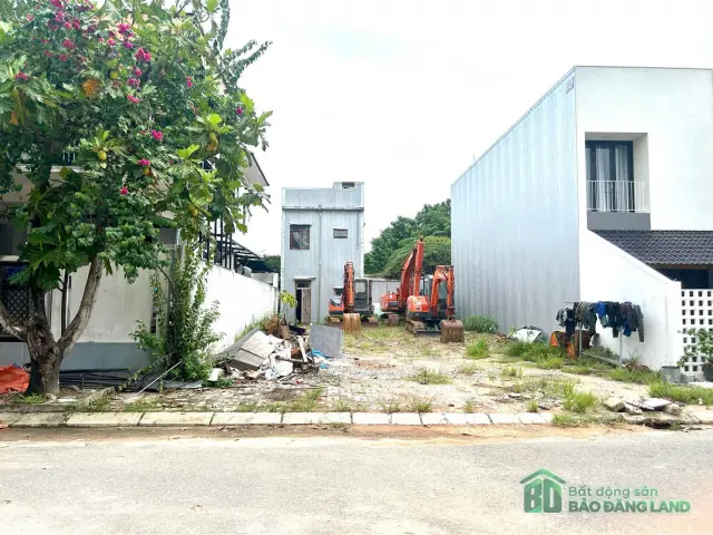 Lô đất trống nhìn từ phía đường với cây hoa đỏ và xe đào bên trong