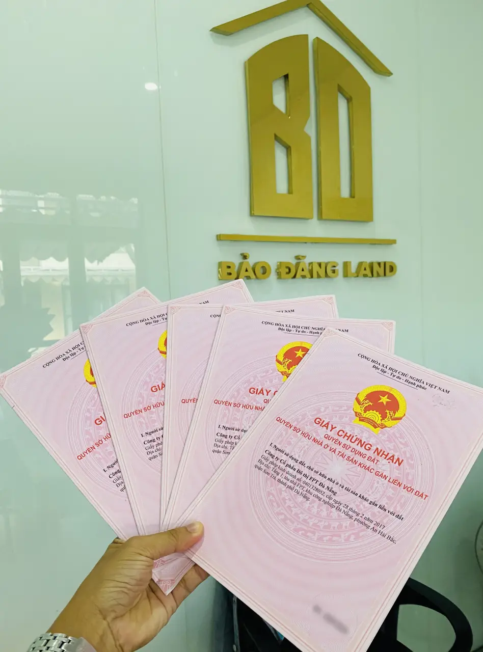 Tập sổ đỏ cầm trên tay trước bảng hiệu logo công ty Bảo Đăng Land.